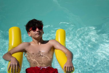 Genç çocuk yaz tatilinin tadını çıkarıyor. Sarı şişme yatakta yüzüyor. Fotokopi alanı olan turkuaz havuz suyunda.