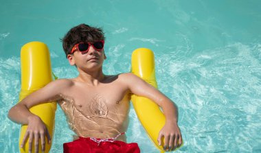Genç çocuk yaz tatilinin tadını çıkarıyor, turkuaz yüzme havuzunda sarı şişme bir yatakta yüzüyor.