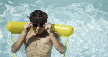 Genç adam yaz tatilinin tadını çıkarıyor, turkuaz havuzdaki sarı şişme yatakta yüzüyor, kırmızı güneş gözlüklerini ayarlıyor, fotokopi alanı ile.