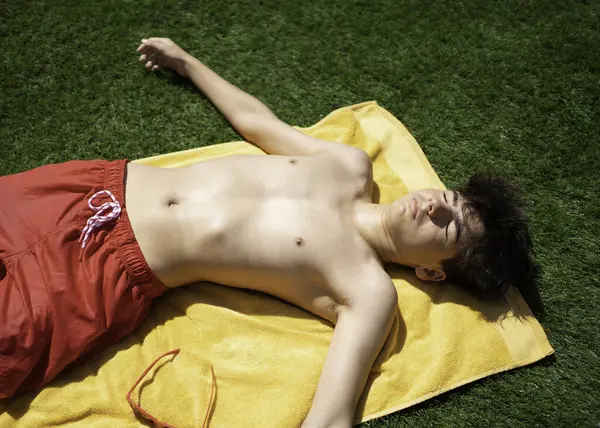 Gözleri kapalı genç bir adam güneşin altında güneşlenen bir havluda dinleniyor.