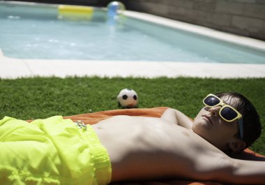 Küçük çocuk havuz kenarında güneşleniyor, güneş gözlüğü takıyor ve yaz tatilinin tadını çıkarıyor.