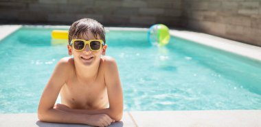 Güneşli bir günde fotokopi çekilen havuz başında dinlenen, güneş gözlüklü gülümseyen çocuğun ön görüntüsü.