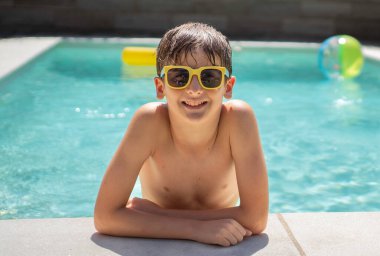 Güneş gözlüklü gülümseyen genç bir çocuğun portresi havuzun kenarında dinleniyor.
