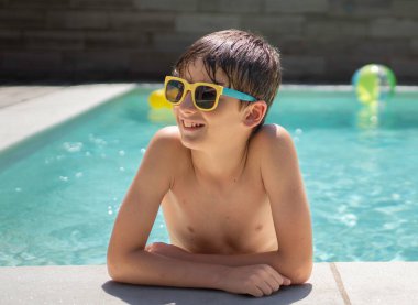Güneş gözlüklü gülümseyen genç bir çocuğun portresi havuz kenarında dinleniyor ve başka tarafa bakıyor.