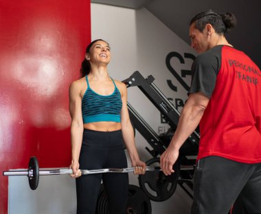 Modern bir spor salonunda kişisel antrenörünün yardımıyla ağırlık kaldıran mutlu sporcu kadın.