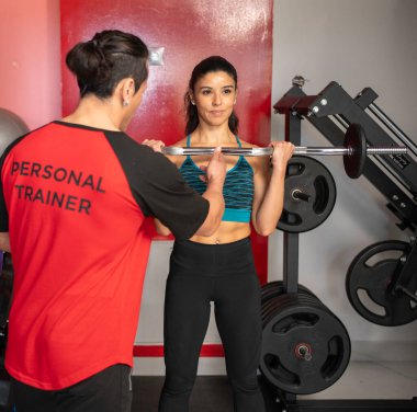 Modern bir spor salonunda ağırlık kaldıran bir kadına yardım eden kişisel antrenör.