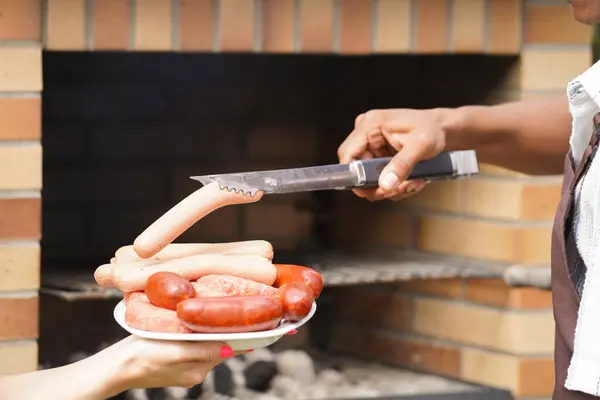 Barbekü partisinde spatulayla ızgaraya koymak için bir tabaktan sosis alırken yaklaş.