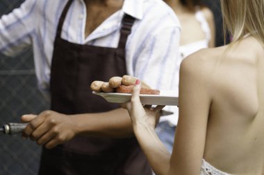 Barbekü partisinde elinde bir tabak ızgara sosis ve hamburger tutan bir kadın, aşçı erkek arkada ızgara yapıyor.