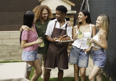 Neşeli arkadaşlar barbekü yapıyorlar, ızgara yemek ve içecekler tutuyorlar, arka bahçede birlikte eğleniyorlar.
