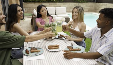 Bir grup çok ırklı arkadaş barbekü partisinde limonata içip ızgara et ve sosis yiyorlar.