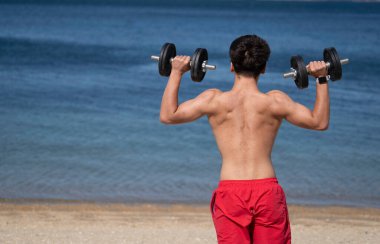 Sahilde spor yapan genç bir sporcunun arka görüntüsü ağırlık kaldırıyor ve forma olan bağlılığını ve gücünü gösteriyor.