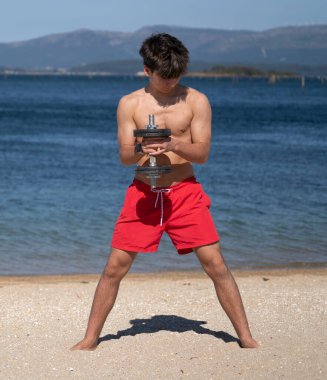 Tişörtsüz genç adam güneşli bir sahilde ağırlık kaldırıyor yaz tatilinde sağlıklı ve aktif bir yaşam tarzının tadını çıkarıyor.