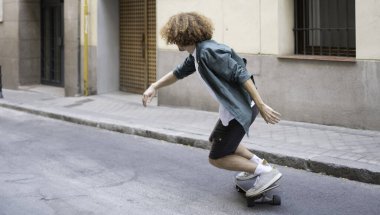 Kentsel caddede kıvırcık saçlı kaykaylı genç bir adam.