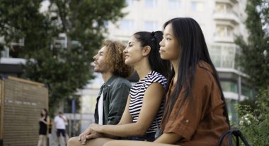 Üç genç çoklu etnik öğrenci şehir ortamında oturuyorlar, uzaktaki bir şeye bakıyorlar.