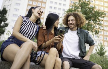Üç neşeli çoklu etnik arkadaş dışarıda birlikte oturuyorlar, akıllı telefon kullanırken birlikte vakit geçiriyorlar.