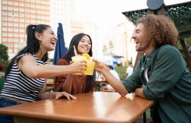 Çok ırklı mutlu arkadaşlar, açık bir kafede meyveli içeceklerin tadını çıkarırken birlikte gülüp kadeh kaldırıyorlar.