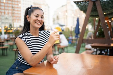 Mutlu genç Latin kadın gülümsüyor ve açık hava yaz festivalinde ferahlatıcı bir içki tutuyor, fotokopi uzayı ile canlı atmosferin tadını çıkarıyor.