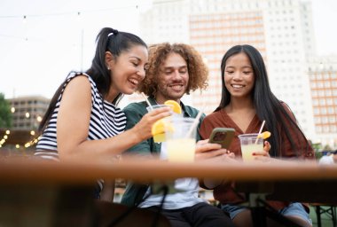 Üç mutlu arkadaş canlı bir açık hava kafedeki akıllı bir telefondan içecek ve sosyal medyaya göz gezdiriyorlar.