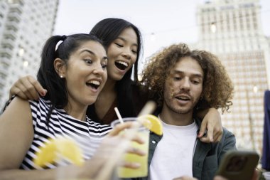 Çatıdaki bir barda kokteyl içerken akıllı telefondan bir şeyler izleyen heyecanlı arkadaşlar.