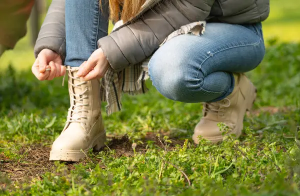 Kotlu ve gri ceketli genç bir kadının, bej çizmelerinin bağcıklarını bağlarken ormanın çimlerinde çömelirken