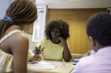 Siyahi öğretmen iki öğrenciyle aynı sınıfta akademik materyalleri tartışıyor.