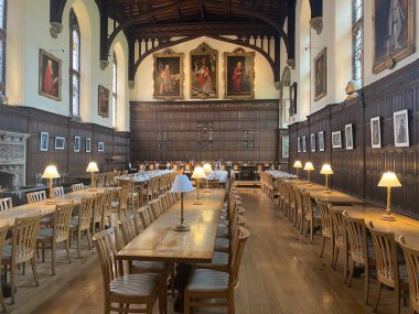 Oxford Magdalen Koleji 'nin tarihi yemek salonu. Uzun ahşap masalar, sandalyeler ve masa lambaları. Süslü ahşap paneller, kemerli tavan. Rönesans resimleri duvarları süslüyor. Büyük, akademik atmosfer.