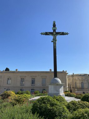 Historic crucifix in Montpellier, France, standing against azure sky near Place Royal du Peyrou. Elegant French classical buildings and manicured Mediterranean garden create stunning composition. clipart