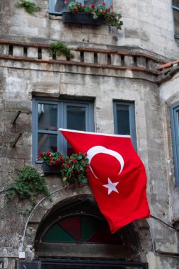 Türk bayrağının öne çıktığı tarihi taş bina cephesi. Pencere kutularında kırmızı çiçeklerle süslenmiş mavi çerçeveli pencereler. Kemerli kapı aralığı ve kırsal mimari detaylar