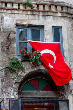 Turkish flag proudly displayed on historic stone building facade with blue-framed windows, decorative arch doorway, and vibrant red flowers in window boxes. Rustic architectural details clipart