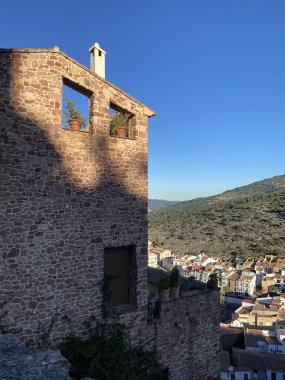 Historic stone tower overlooks Mediterranean village. Rustic architecture with terracotta pots in arched windows. Dramatic mountain landscape and traditional townscape below clear blue sky. clipart