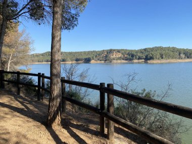 Saf ahşap çitleri ve uzun çam ağacı olan sakin göl manzaralı bir yer. Açık mavi su, parlak gökyüzünün altında ormanlık tepelerle buluşur. Doğa yolu manzaralı bir bakış sunuyor..