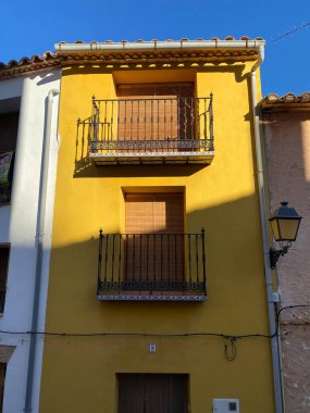 Geleneksel İspanyol konak cephesinde parlak sarı duvarlar, iki süslü demir balkon, terasotta çatı kiremitleri ve parlak mavi gökyüzüne karşı ahşap panjurlar var..
