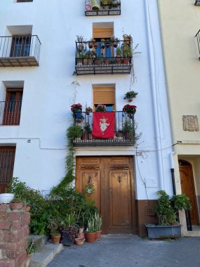 Ahşap kapılı Büyüleyici Akdeniz bina cephesi, süslü balkonlar, saksı bitkileri, çiçekler ve belirgin kırmızı bir afiş. Beyaz duvarlar ve kırsal elementler misafirperver bir atmosfer yaratıyor