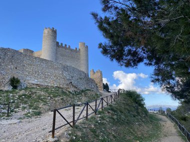 Tepesinde silindirik kuleleri olan ortaçağ taş kalesi. Antik tahkimat duvarları, canlı mavi gökyüzüne karşı. Ahşap korkuluklu dolambaçlı yol girişe çıkıyor. Çam ağaçları tarihi bir dönüm noktası.