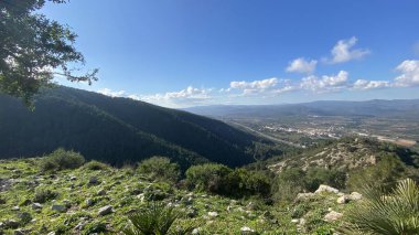  Tepeleri yuvarlanan panoramik dağ manzarası, vadideki uzak kasaba. Engebeli arazide yeşil bitki örtüsü ve dağınık kayalar var. Beyaz bulutlu parlak mavi gökyüzü dramatik bir manzara yaratır..