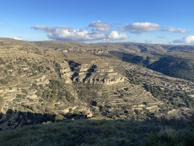 Parlak mavi gökyüzünün altında teraslı yamaçlar ve kireçtaşı kayalıkları ve dağınık beyaz bulutlarla dramatik bir dağ manzarası. Arid Akdeniz arazisi doğal aşınma şekillerini gösteriyor.