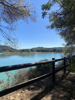 Tahta çitlerle çevrili yürüyüş parkurundan görülen sakin turkuaz göl. Açık mavi gökyüzü, sakin suları çevreleyen zümrüt ormanlarıyla çelişiyor. Tepeler ufku çerçeveler. Mükemmel sakin doğa manzarası..