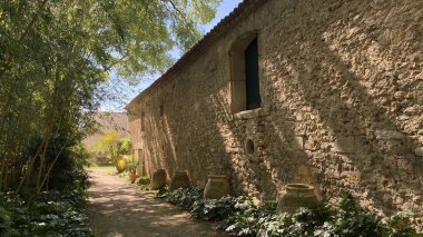 Yıpranmış duvarları olan taştan bir çiftlik evi. Dar toprak patika, antik binanın yanında. Çamur tencereleri ve yemyeşil alanlar parlak güneş ışığı altında Akdeniz kırsal atmosferini oluşturur.