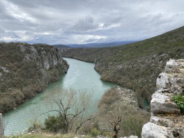 Engebeli kireçtaşı kanyonunu oyan yeşim taşı yeşili Cetina Nehri 'nin göz kamaştırıcı hava manzarası. Uzak dağların üzerinde dramatik bulutlu gökyüzü beliriyor. Nadir bitki örtüsü dik kayalık yamaçlara tutunur..