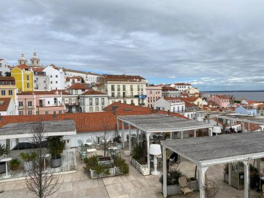Lizbon 'un Alfama bölgesinin panoramik manzarası renkli binalar ve terracotta çatıları. Ön planda beyaz pergolalı çatı terası var. Arkaplanda Tagus Nehri ile tarihi şehir manzarası üzerinde bulutlu bir gökyüzü.