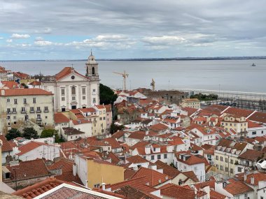 Lizbon 'un tarihi Alfama bölgesinin panoramik manzarası. Karakteristik terracotta çatıları var. Çan kulesi olan beyaz kilise göze çarpıyor. Tagus Nehri ufuk boyunca uzanıyor parçalı bulutlu gökyüzünün altında.