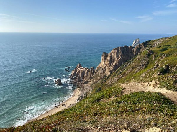 Драматическое португальское побережье с возвышающимися морскими стеками и бирюзовыми водами. Крепкие скалы падают на уединенный пляж внизу. Роскошная прибрежная растительность.