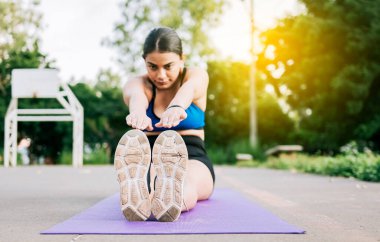 Sporcu kadın parkta bacak esneme hareketleri yapıyor. Parkta bacaklarını esneten bir paspasın üzerinde oturan sporcu kız, parkta bir paspasın üzerinde oturan sportif kız.