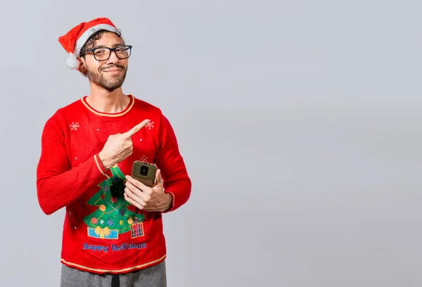 Christmas man holding phone pointing to the side. Smiling young man in christmas hat with cellphone pointing at promo. Christmas man concept with cell phone pointing to the side