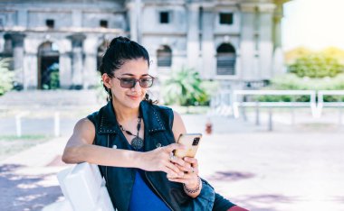 Urban style girl sitting on a bench with her cell phone, An attractive girl sitting on the cell phone smiling, Girl sitting on a bench texting on her cell phone.