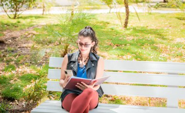 A girl sitting taking notes with a notebook and cell phone, Woman sitting taking notes with her cell phone, Latin girl using her cell phone in a park
