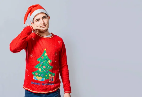 Man in christmas clothes imitating a phone conversation, Young latin man in christmas hat making a call gesture isolated. People in Christmas clothes making a call gesture with their fingers