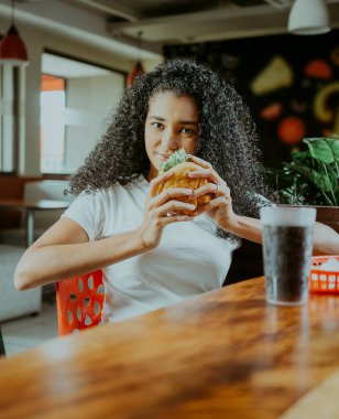 Güzel Latin kız bir restoranda lezzetli bir hamburgerin tadını çıkarıyor. Restoranda elinde hamburger tutan afro bir kızın portresi..