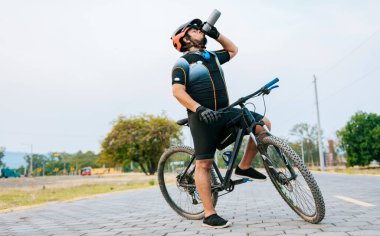 Tombul bisikletçi bisikletiyle su içiyor susamış bisikletçi dışarıda su içiyor. Dışarıda su içen yorgun bir bisikletçi.