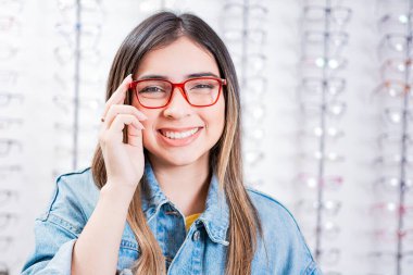 Optik mağazasının arka planında gözlüklü mutlu genç kız. Optik mağazada mutlu kadın modellik gözlüğü